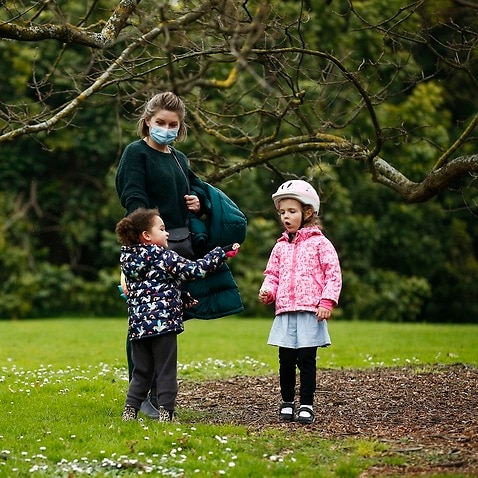 A woman wears a mask as two young children play in the Botanic Gardens in Melbourne