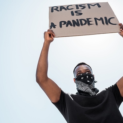 low angle view of african american man with scarf on face holding placard with racism is a pandemic lettering against blue sky