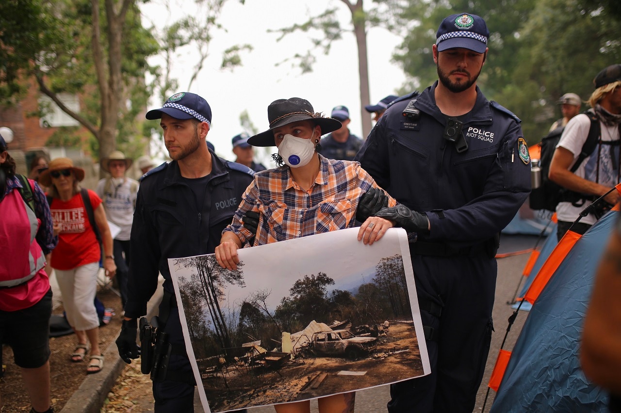 A protester holding a photograph of property destroyed by bushfire is arrested outside of Kirribilli House. A protester holding a photograph of property destroyed by bushfire is arrested outside of Kirribilli House.