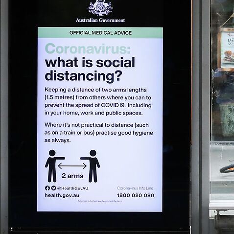 A commuter sits on a tram bench next to an LCD screen with an Australian Government directive regarding Coronavirus and social distancing