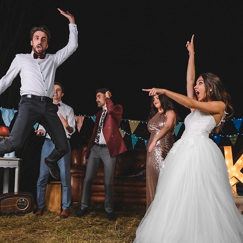 Surprised bride looking at man jumping on a night field party with friends