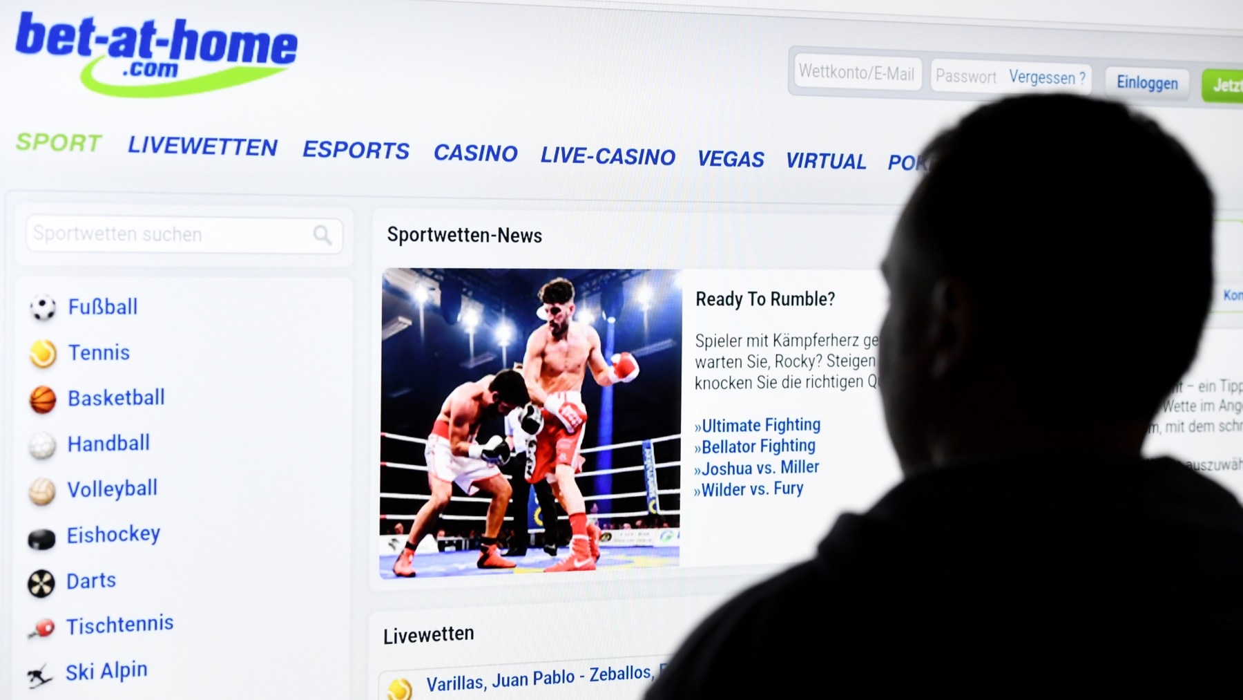 A man stands in front of a screen with a page of a sports betting provider (bet-at-home)