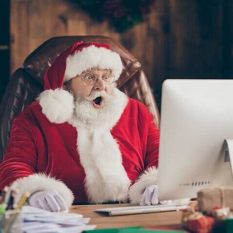 Photo of amazed santa claus sit table desk work computer impressed many wish gift list letters in house idnoors with evergreen christmas x-mas tree ornament decoration wear cap headwear