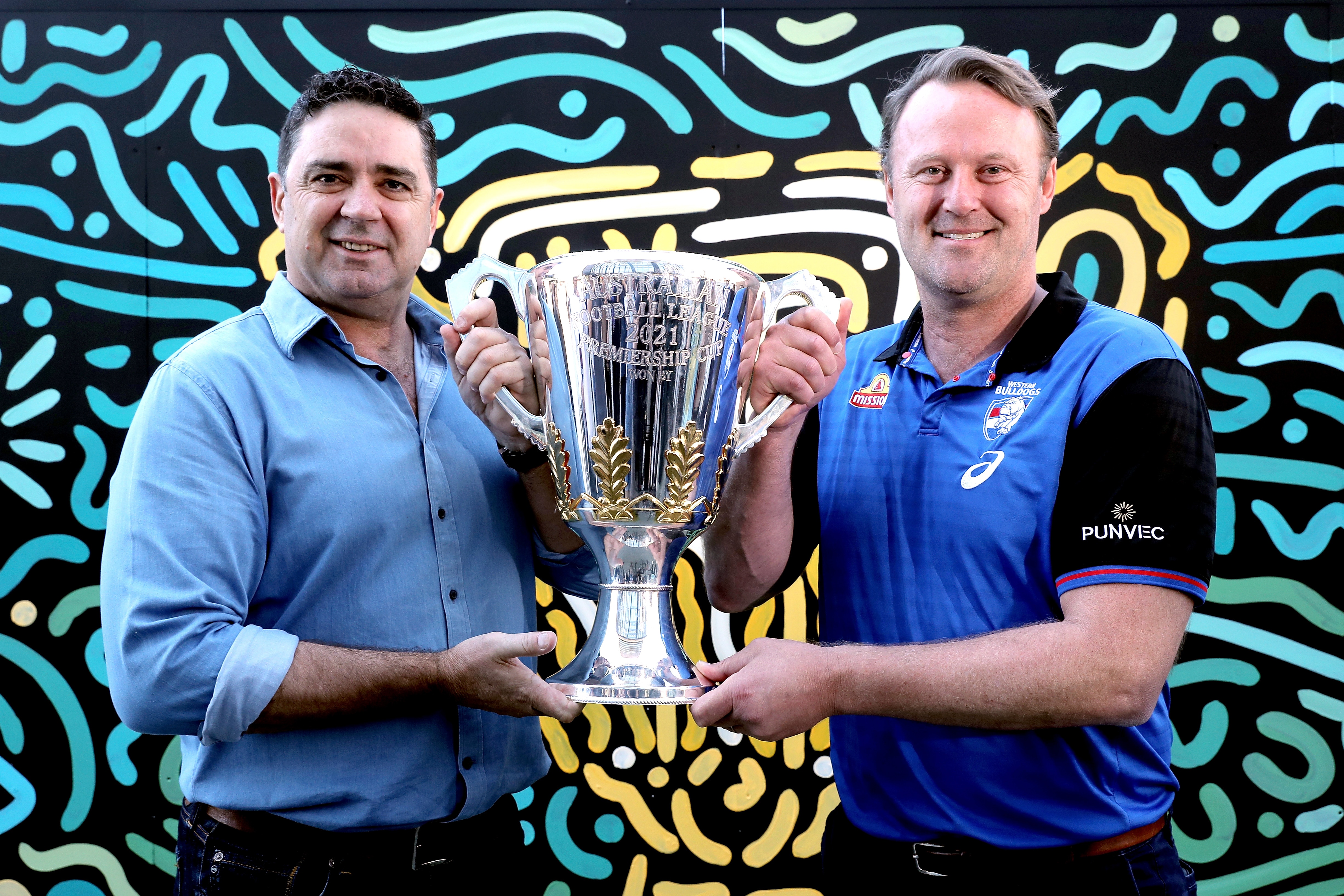 2021 AFL Premiership Cup presenters Garry Lyon and Chris Grant pose for a photograph in front of a mural by local Perth artist Kamsani Kambarni Bin Salleh.