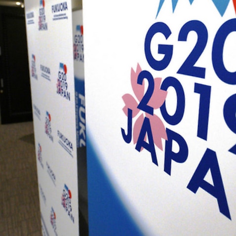 Staff members stand near the emblem of G20 2019 Japan at the entrance of the press centre